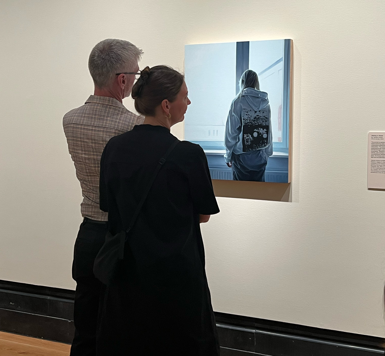 Two gallery visitors standing before the painting Ukranian girl, contemplating the composition of the young woman viewed from behind by the window within the institutional gallery setting