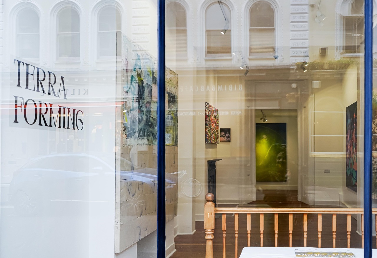 Night-time view from outside the gallery at Museum Street, Bloomsbury, 2025. TERRA FORMING text on the glass overlaps with reflections of the Victorian building opposite; artworks from the Terraforming exhibition are visible inside.