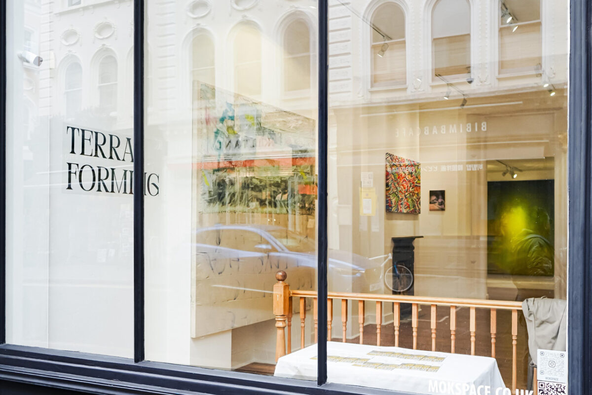 Vista diurna desde la calle de la galería en Museum Street, Bloomsbury, durante Terraforming, 2025. El título TERRA FORMING aparece en el vitral junto a un código QR; al interior se aprecia la disposición de las obras en el espacio expositivo.