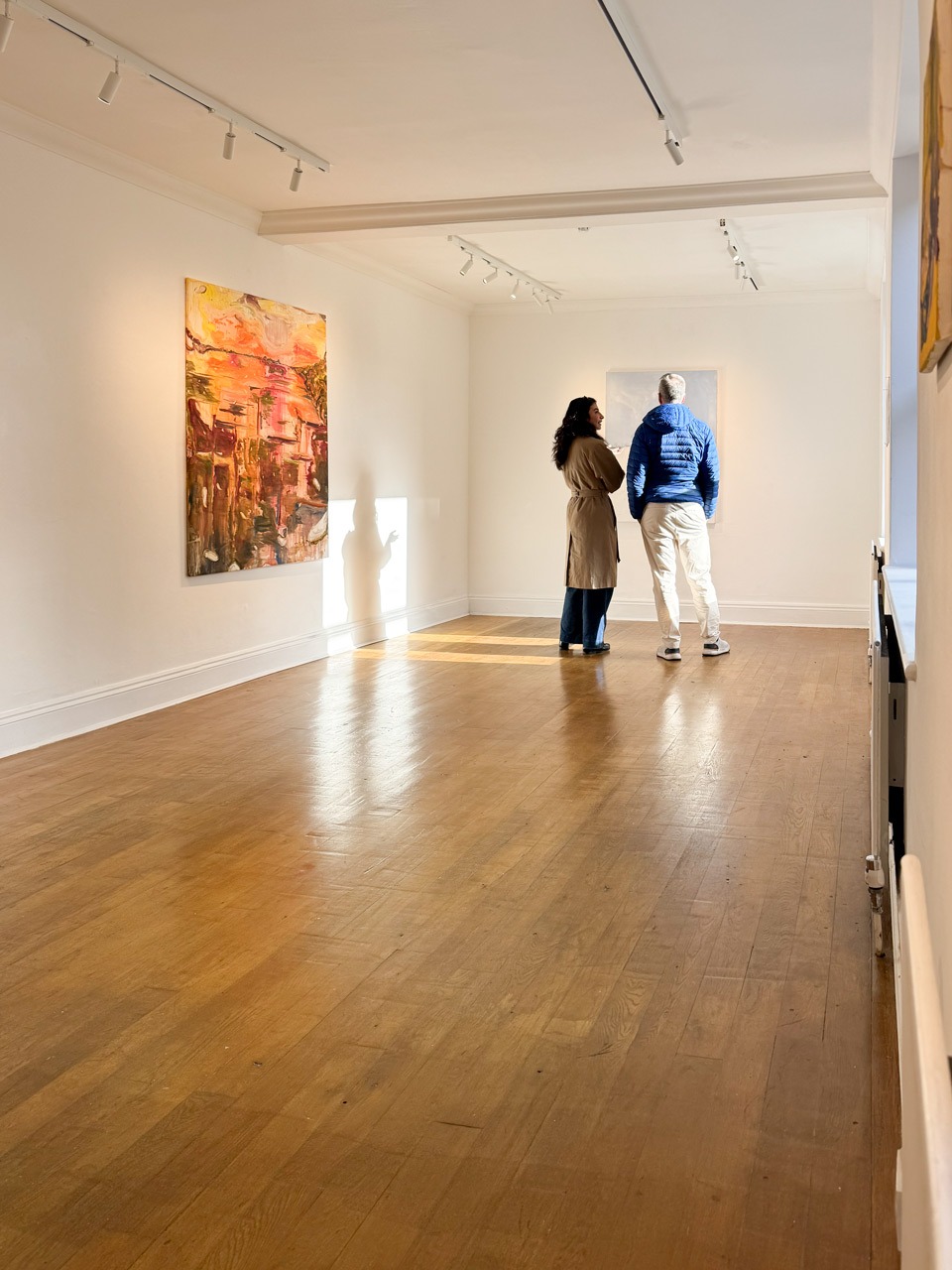 Two visitors viewing large-scale paintings in the main room at Pipeline Gallery during What we take, March 2026. Expressive works in warm tones hang on white walls under track lighting.
