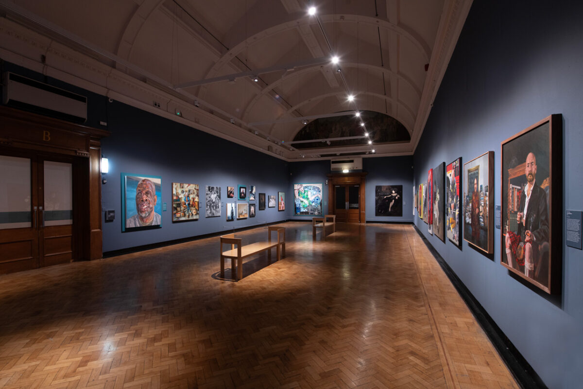 Second room of the exhibition at the Laing Art Gallery, seen from the wooden doors, portraits on blue-grey walls with benches under directional lighting