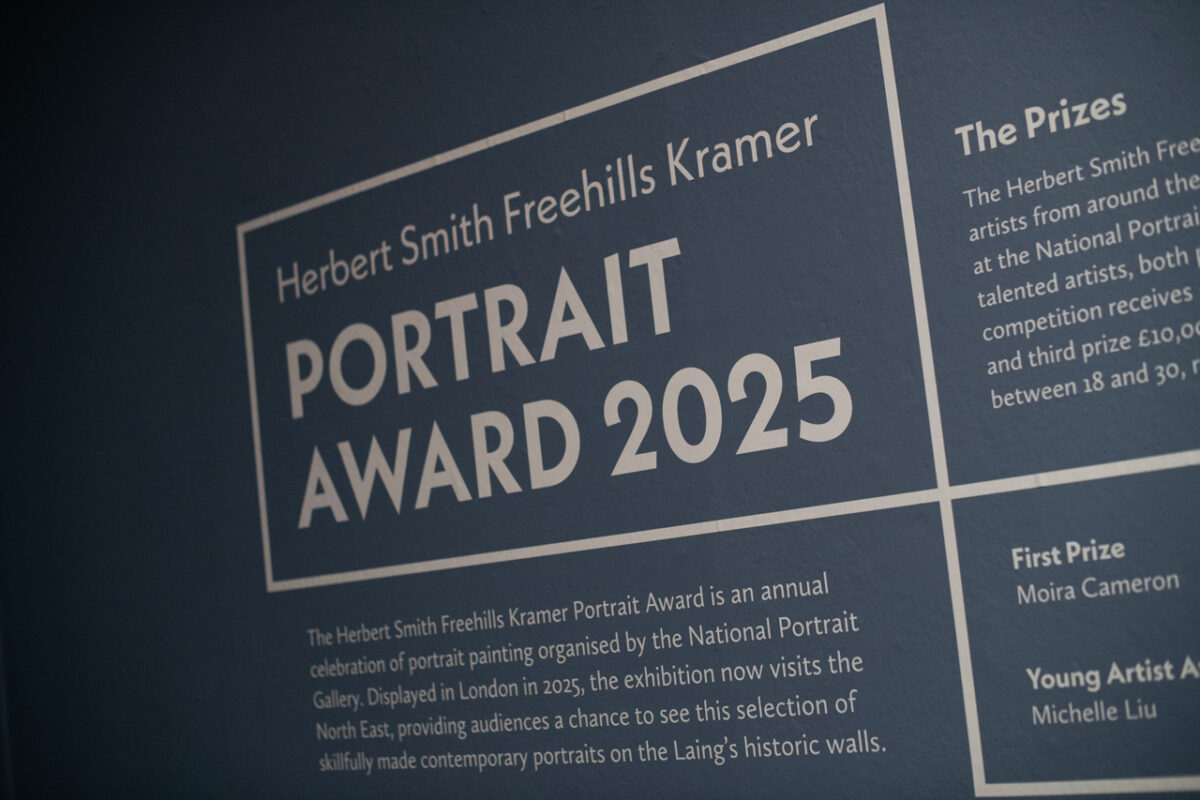 Detail of the Herbert Smith Freehills Kramer Portrait Award 2025 wall text, photographed at an angle against the blue wall of the Laing Art Gallery