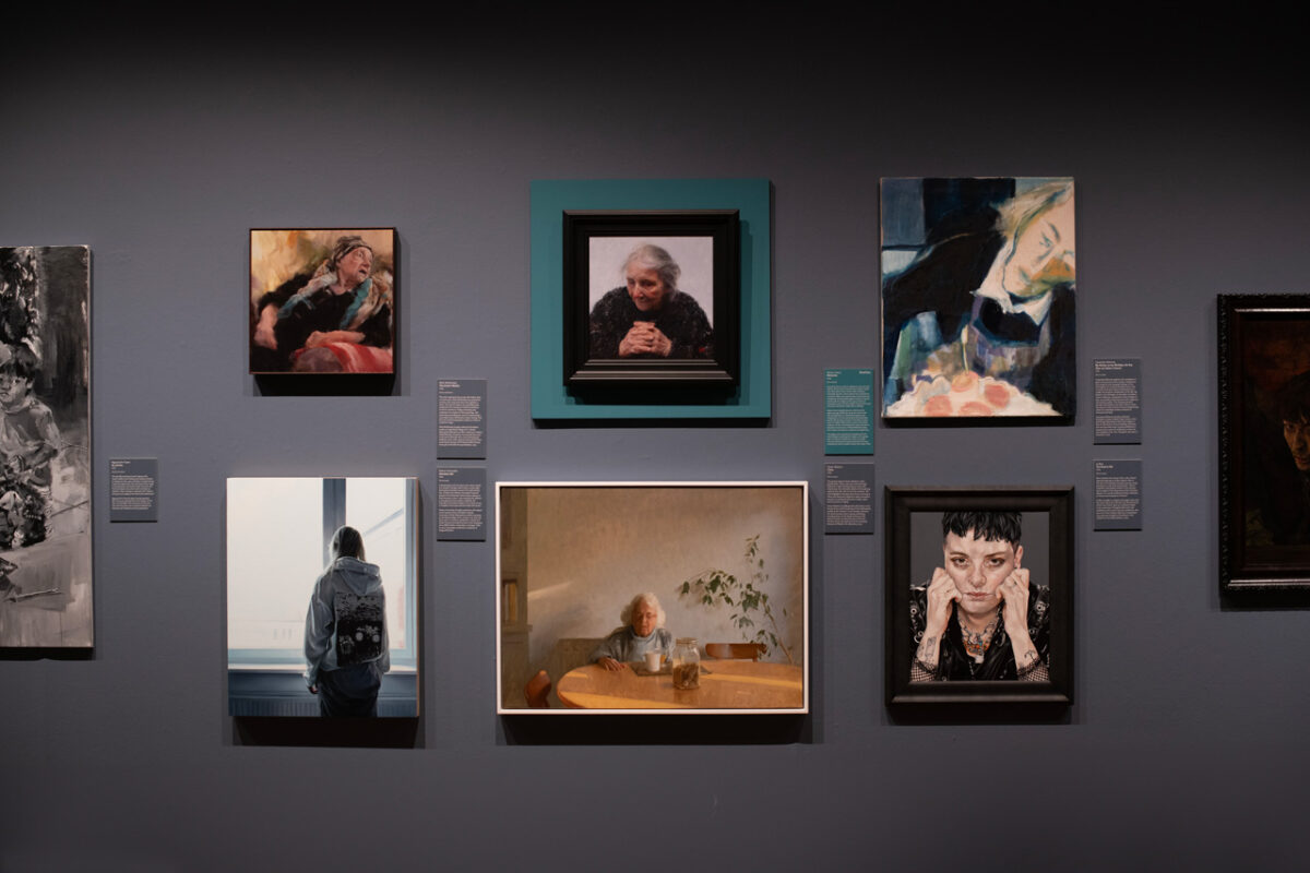 Exhibition wall with eight portraits; lower left, Ukranian girl by Nelson Hernández, a young woman seen from behind in a grey hoodie facing a window